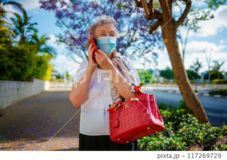 An 80-year-old woman, in a white blouse and face mask, smiles while talking on her red phone and holding a red handbag under a jacaranda tree on a city sidewalk. 117269729