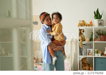 Woman lovingly holding her child in a cozy, well-decorated living space surrounded by toys and bookshelves, featuring plants and stuffed animals on shelves 117279260