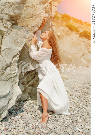 A woman in a white dress is standing on a rocky beach. Concept of serenity and tranquility, as the woman is enjoying the natural beauty of the beach. 117279737