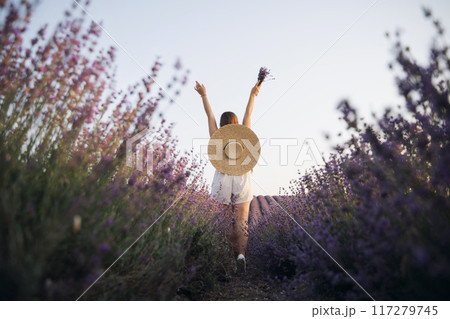 A woman is walking through a field of purple flowers with a straw hat on 117279745