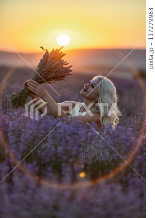 Blonde woman poses in lavender field at sunset. Happy woman in white dress holds lavender bouquet. Aromatherapy concept, lavender oil, photo session in lavender Blonde woman poses in lavender field at sunset. Happy woman in white dress holds lavender bouquet. Aromatherapy concept, lavender oil, photo session in lavender 117279963