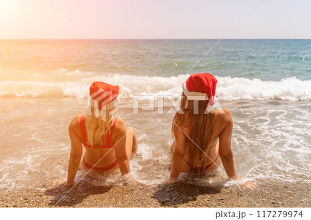 Women Santa hats ocean play. Seaside, beach daytime, enjoying beach fun. Two women in red swimsuits and Santa hats are enjoying themselves in the ocean waves. Women Santa hats ocean play. Seaside, beach daytime, enjoying beach fun. Two women in red swimsuits and Santa hats are enjoying themselves in the ocean waves. 117279974