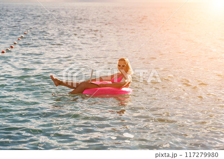 Woman works on laptop in sea. Freelancer, blond woman in sunglases floating on an inflatable big pink donut with a laptop in the sea at sunset. People summer vacation rest lifestyle concept. Woman works on laptop in sea. Freelancer, blond woman in sunglases floating on an inflatable big pink donut with a laptop in the sea at sunset. People summer vacation rest lifestyle concept. 117279980