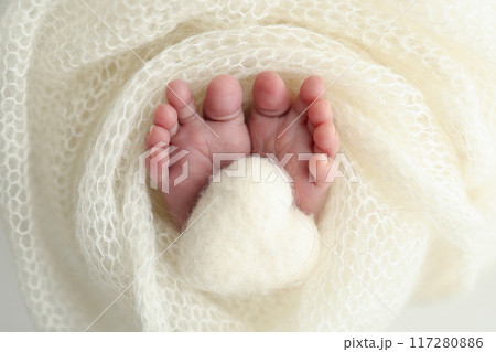 The tiny foot of a newborn baby. Soft feet of a new born in a white wool blanket. Close up of toes, heels and feet of a newborn. Knitted white heart in the legs of a baby. Macro photography.  117280886