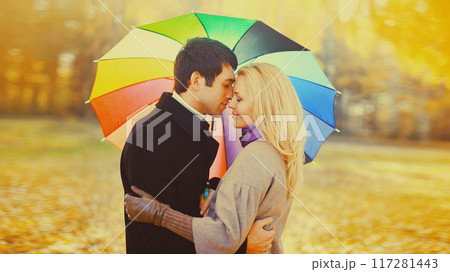 Happy loving young couple kissing hugging under colorful umbrella in autumn park on warm sunny day 117281443