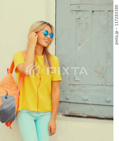 stylish modern blonde young woman listening to music in headphones, glasses in the city 117283380