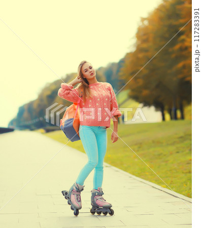 Stylish blonde young woman on roller skates with backpack posing in city park 117283391
