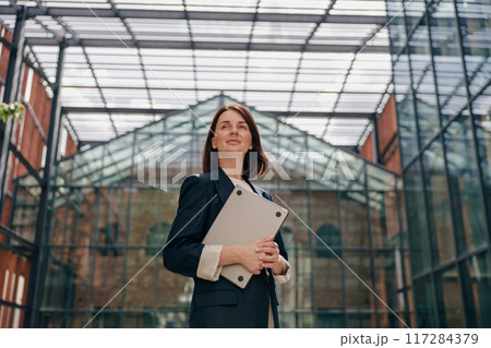 A confident businesswoman is standing in modern glass architecture, holding documents 117284379