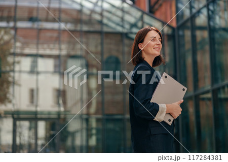 A successful businesswoman confidently holds a laptop in front of a contemporary office building 117284381