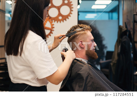 Red-haired bearded man sitting in barbershop and female barber making hairdo to guest 117284551