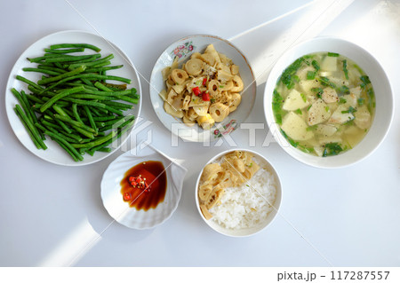 Vietnamese meal with steamed rice, tofu and vegetable soup, stir-fried green beans, bamboo shoots, and chili sauce on a white table, illuminated by natural light from a window. Vietnamese meal with steamed rice, tofu and vegetable soup, stir-fried green beans, bamboo shoots, and chili sauce on a white table, illuminated by natural light from a window. 117287557
