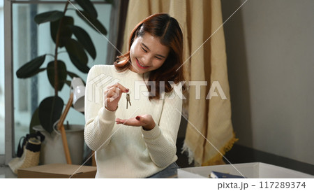 Happy young woman holding keys sitting in her new home with moving boxes. Buying or renting property concept Happy young woman holding keys sitting in her new home with moving boxes. Buying or renting property concept 117289374