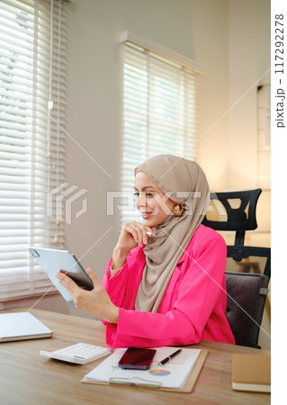 A woman wearing a pink jacket is sitting at a desk with a tablet in front of her 117292278