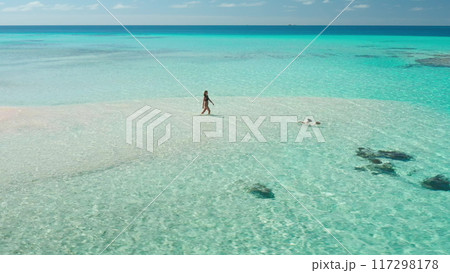 Breathtaking aerial view of a young woman enjoying a leisurely walk on a pristine sandbank, her friend swimming in the crystal-clear turquoise waters of the maldives 117298178