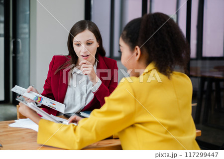 Two professional women engaged in a collaborative discussion in a modern office, reviewing documents and sharing ideas. 117299447