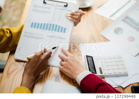 Colleagues discussing financial data and charts during a business meeting, with a calculator and documents on a wooden desk. Colleagues discussing financial data and charts during a business meeting, with a calculator and documents on a wooden desk. 117299448