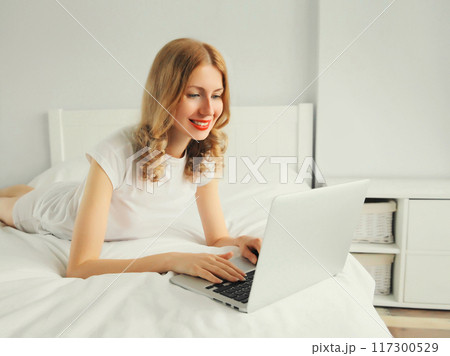 Portrait of smiling young woman working with laptop on the bed in white room at home 117300529
