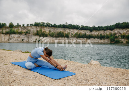 A woman is seen practicing yoga surrounded by the serene and picturesque environment of a lake A woman is seen practicing yoga surrounded by the serene and picturesque environment of a lake 117300926