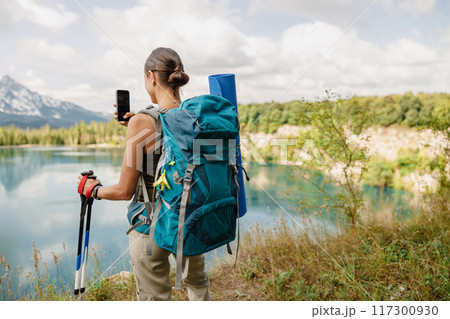 A hiker is taking beautiful photos by a scenic lake while wearing a stylish backpack 117300930