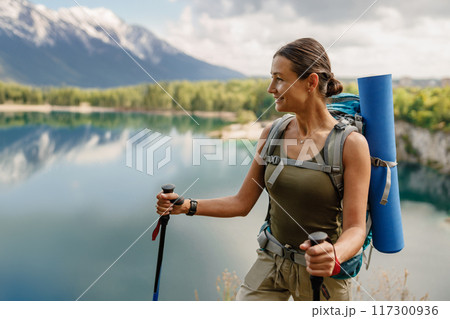 A young woman is hiking joyfully near beautiful mountains alongside a serene lake A young woman is hiking joyfully near beautiful mountains alongside a serene lake 117300936