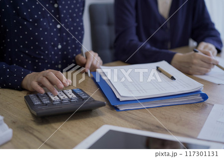 Accountants or auditors reviewing financial documents while sitting at the wooden desk in office. Professionals analyzing paper reports and using calculators, side view. Audit and taxes 117301131