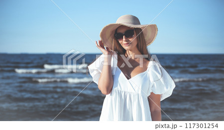 Happy blonde woman is posing on the ocean beach with sunglasses and a hat. Evening sun Happy blonde woman is posing on the ocean beach with sunglasses and a hat. Evening sun 117301254