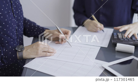 Accountants or auditors reviewing financial documents while sitting at the wooden desk in office. Professionals analyzing paper reports and using calculators, side view. Audit and taxes 117301465