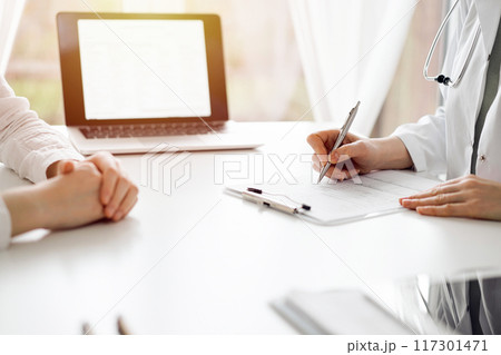 Doctor and patient sitting near each other at the white desk in clinic. Female physician is listening filling up a records form. Medicine concept 117301471