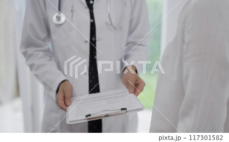 Doctor and a patient discussing something. The female physician, wearing a white medical coat is keeping clipboard with a medical notes besides a young woman during a consultation in the clinic Doctor and a patient discussing something. The female physician, wearing a white medical coat is keeping clipboard with a medical notes besides a young woman during a consultation in the clinic 117301582