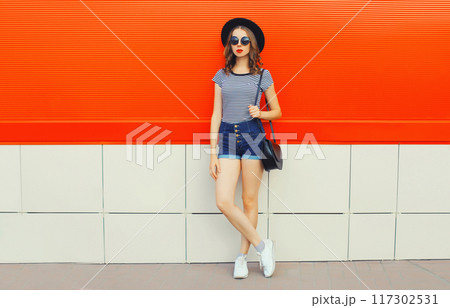 Beautiful woman model posing in black round hat, shorts and handbag in the city on orange background Beautiful woman model posing in black round hat, shorts and handbag in the city on orange background 117302531