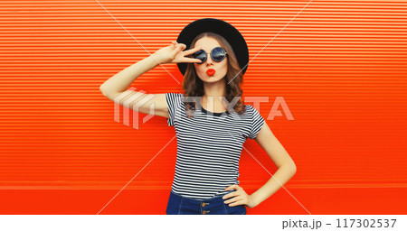 Portrait of beautiful young woman blowing kiss wearing black round hat on orange background Portrait of beautiful young woman blowing kiss wearing black round hat on orange background 117302537