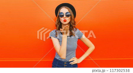 Portrait of beautiful young woman blowing kiss wearing black round hat on orange background 117302538
