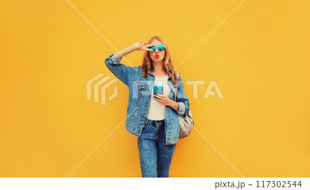 stylish young woman blowing kiss with smartphone posing in jean jacket on yellow background stylish young woman blowing kiss with smartphone posing in jean jacket on yellow background 117302544