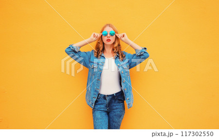 Stylish caucasian blonde young woman posing wearing sunglasses and denim jacket on yellow background Stylish caucasian blonde young woman posing wearing sunglasses and denim jacket on yellow background 117302550