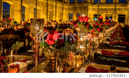 Elegant table set with gold-covered glasses, lighted candles, and red flowers in a historic building. Depth of field used intentionally for effect. Elegant table set with gold-covered glasses, lighted candles, and red flowers in a historic building. Depth of field used intentionally for effect. 117302851