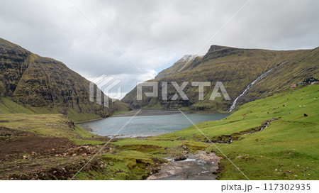 Saksun Lake in the Faroe Islands, framed by lush hills, cliffs, and a waterfall, offers a serene and scenic view from the village of Saksun on Streymoy Island. Saksun Lake in the Faroe Islands, framed by lush hills, cliffs, and a waterfall, offers a serene and scenic view from the village of Saksun on Streymoy Island. 117302935