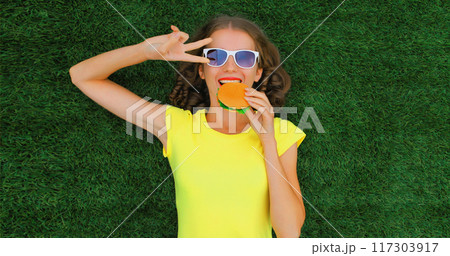 Portrait of happy smiling young woman with big burger fast food lying on the grass in summer park Portrait of happy smiling young woman with big burger fast food lying on the grass in summer park 117303917