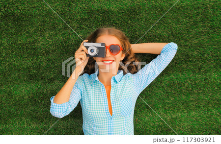 Summer portrait of happy smiling young woman taking pictures on camera lying on the grass in park 117303921