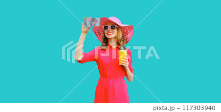 Beautiful happy young woman taking selfie with phone in summer hat, pink dress on blue background 117303940