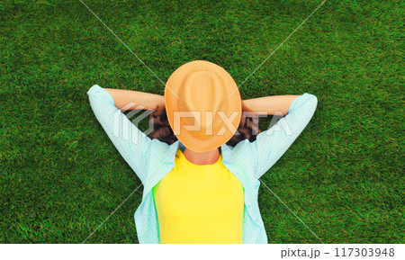 Happy relaxing girl with straw hat covering her face lies on green grass in summer park, top view 117303948