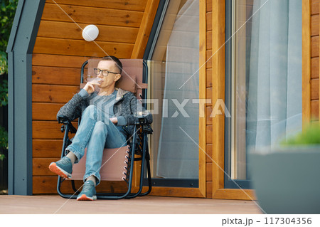 Middle-aged man smoking a cigar while sitting on the terrace of a country house Middle-aged man smoking a cigar while sitting on the terrace of a country house 117304356