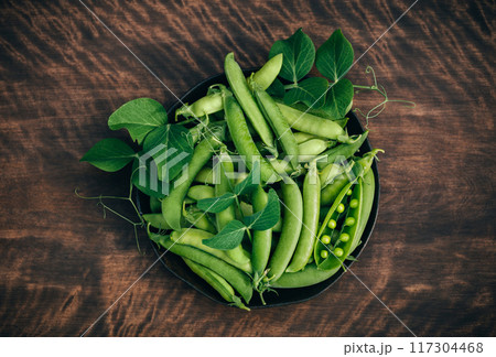 Fresh green peas in a plate on wooden background. Peas with green pods and leaves. Top view, flat lay. 117304468