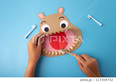 Fun craft project paper-made hippo head with wide-open mouth, cut-out eyes, , hand holding toothbrush, blue background, teaching kids about dental hygiene Fun craft project paper-made hippo head with wide-open mouth, cut-out eyes, , hand holding toothbrush, blue background, teaching kids about dental hygiene 117304732