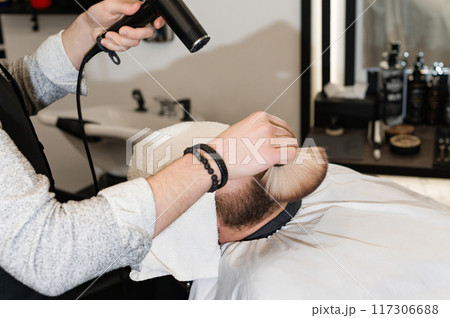 The hairdresser sweeps the hair during the beard trimming with a brush and drying air. The hairdresser sweeps the hair during the beard trimming with a brush and drying air. 117306688