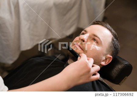 Barber combs beard with comb while shaving Caucasian man Barber combs beard with comb while shaving Caucasian man 117306692