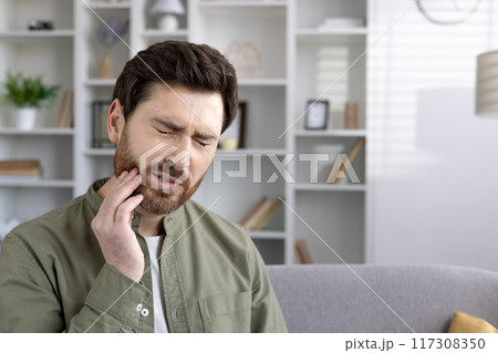Man holding jaw with painful expression experiencing severe toothache in living room. Concept of dental problems, tooth pain, and discomfort. Man holding jaw with painful expression experiencing severe toothache in living room. Concept of dental problems, tooth pain, and discomfort. 117308350