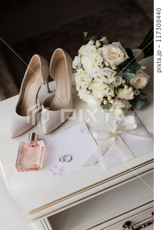 Bride's wedding essentials: heels, bouquet, rings, perfume, and invitation on a white table 117308440