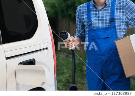 Delivery man in uniform holds electric vehicle charging plug next to electric van 117308987