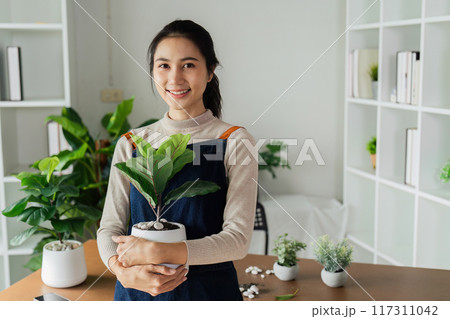 Portrait of a smiling young woman holding potted plant in the house and taking care of the greenery of the house 117311042