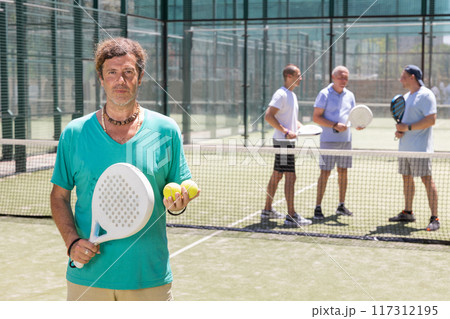 Portrait of man standing on padel tennis court, holding racket and ball 117312195